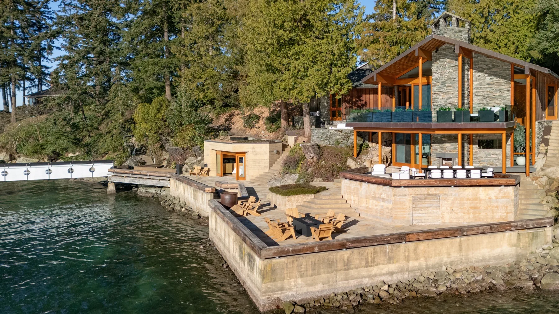 A lakeside house sits beside a stone terrace and outdoor seating.