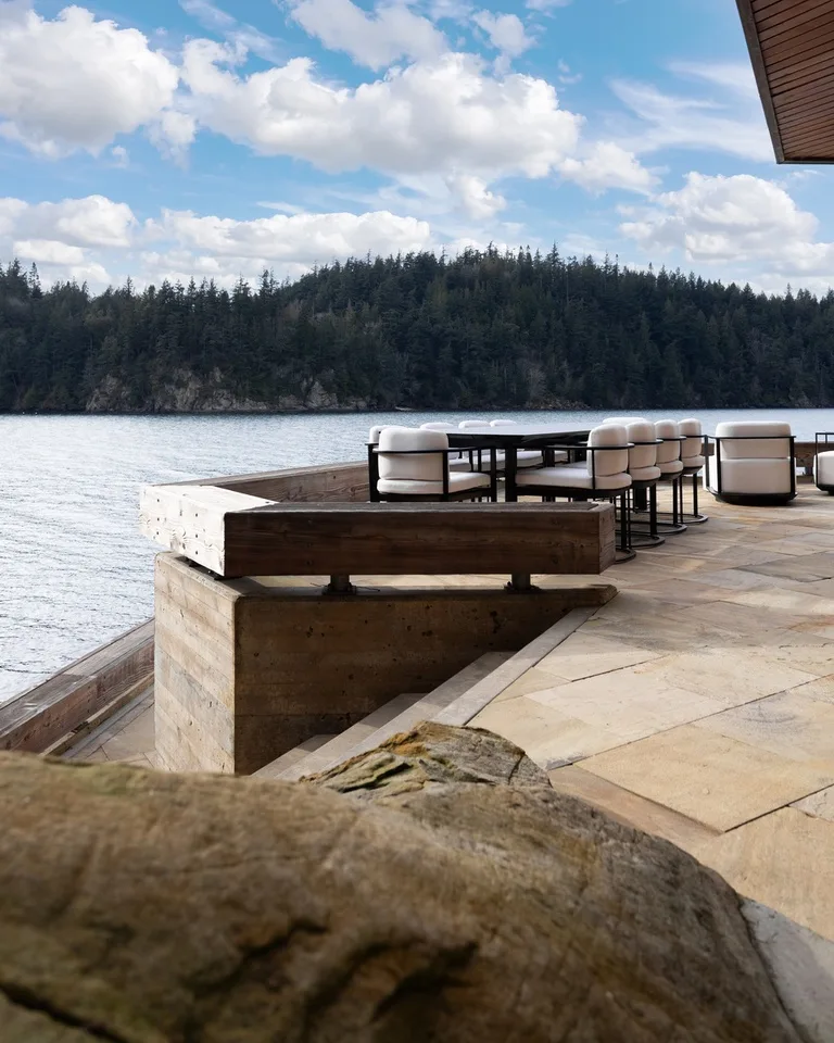 An outdoor dining area beside the water, with tables and seating overlooking a wooded shoreline.