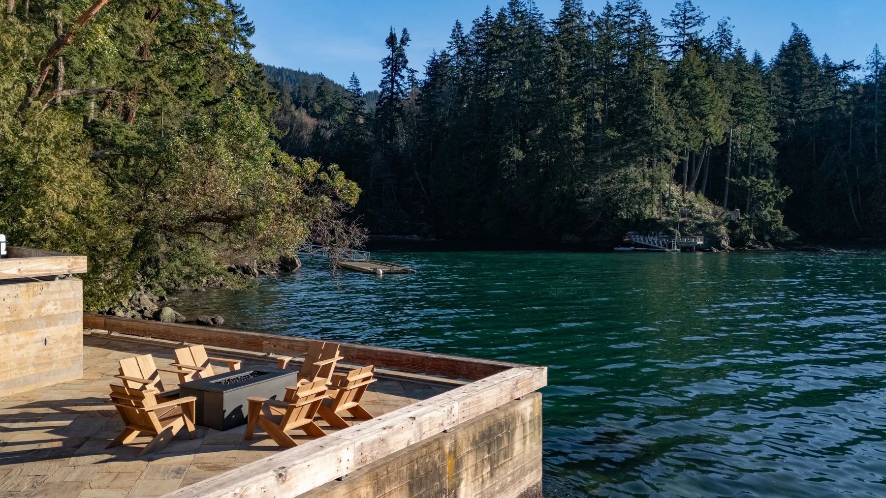 A wooden lakeside deck with seating overlooks green water and a forested shore.