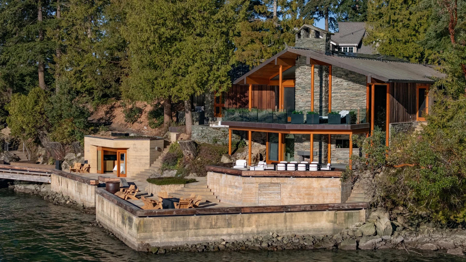 A modern house with a stone exterior sits beside the water among trees.
