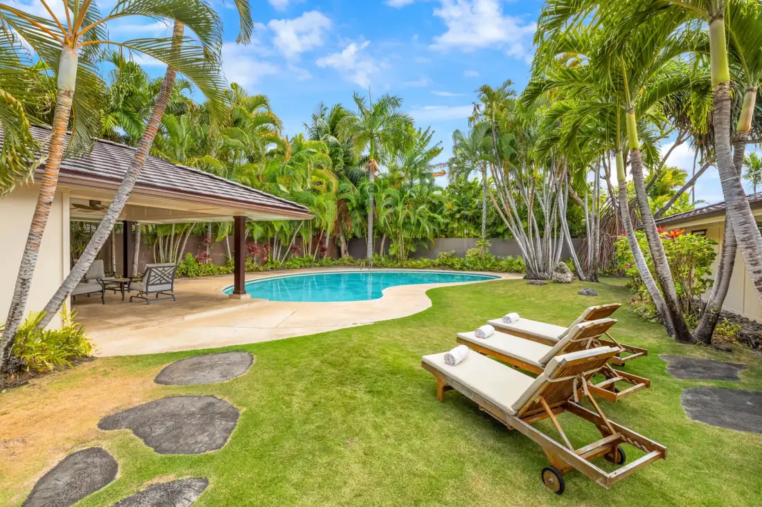A tropical garden surrounds a swimming pool, with lounge chairs on the grass.