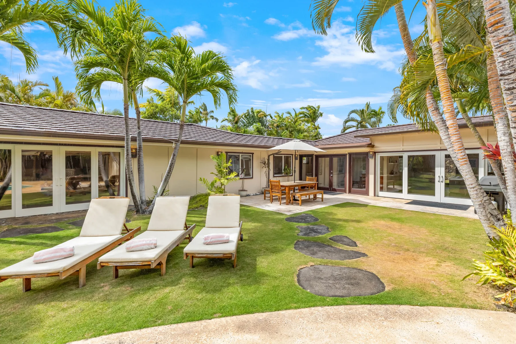 A tropical house opens onto a grassy courtyard with loungers and a dining table.