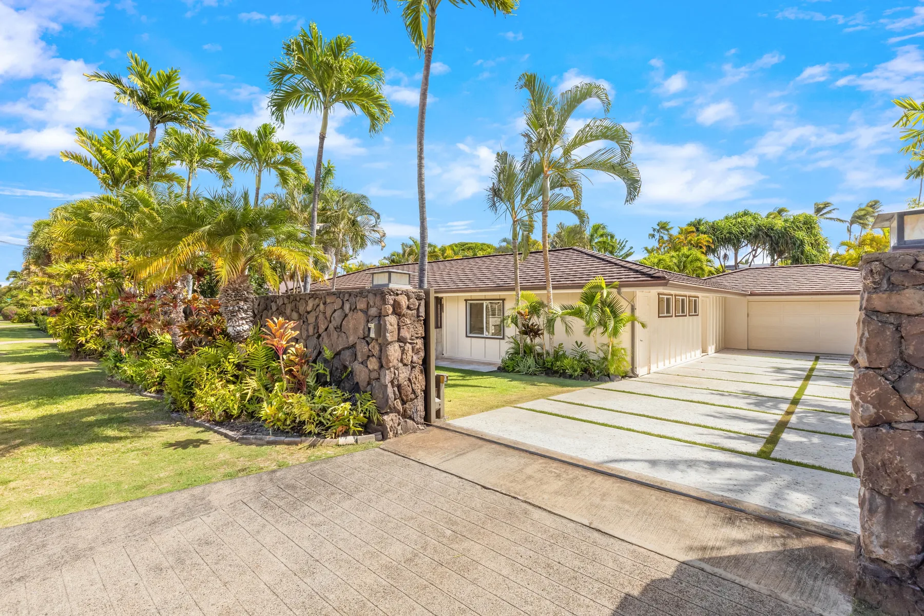 A single-story house stands behind a wide driveway, with palms and tropical landscaping.