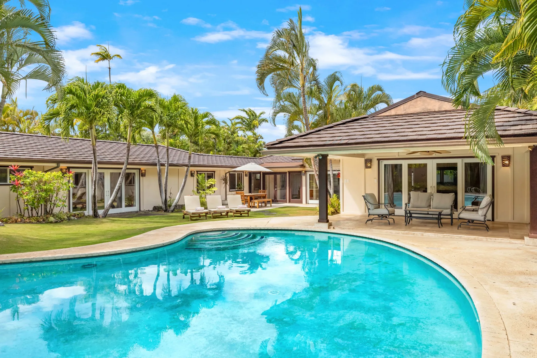A swimming pool curves beside a patio with outdoor seating and palm trees.