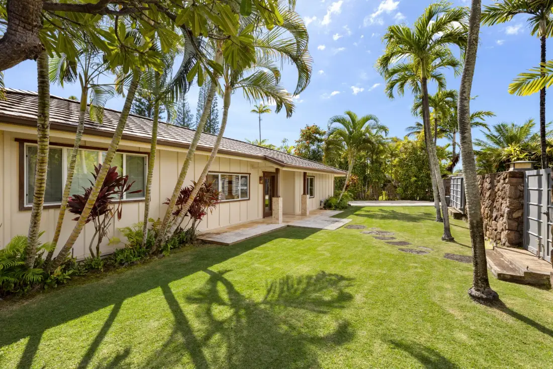 A single-story house faces a sunny lawn with palm trees and tropical plants.