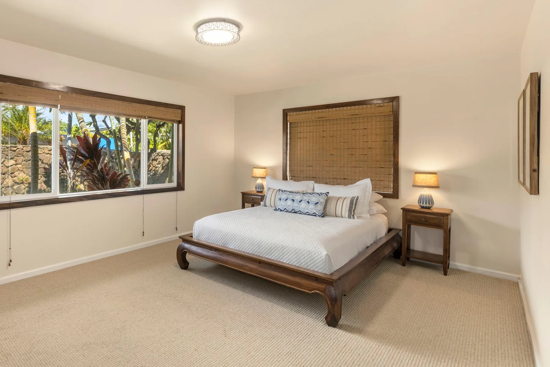 A bedroom with a bed, bedside tables, and windows overlooking tropical greenery.