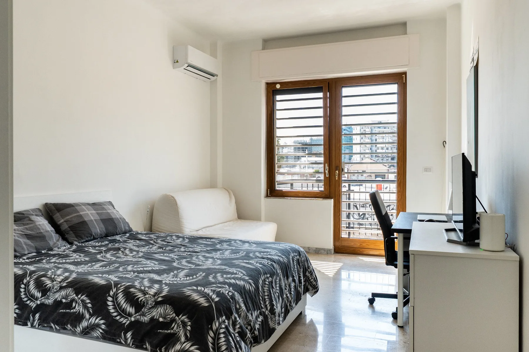 A bright bedroom with a bed, desk, and large windows overlooking nearby buildings.