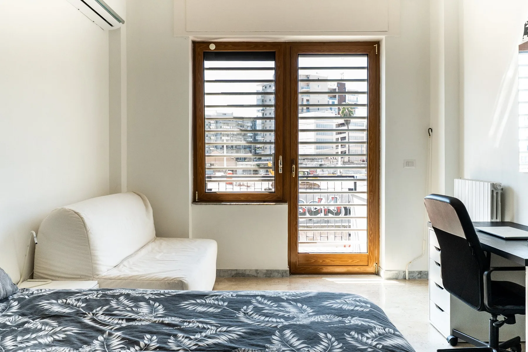 A bright bedroom with a bed, seating, and a desk beside tall windows.