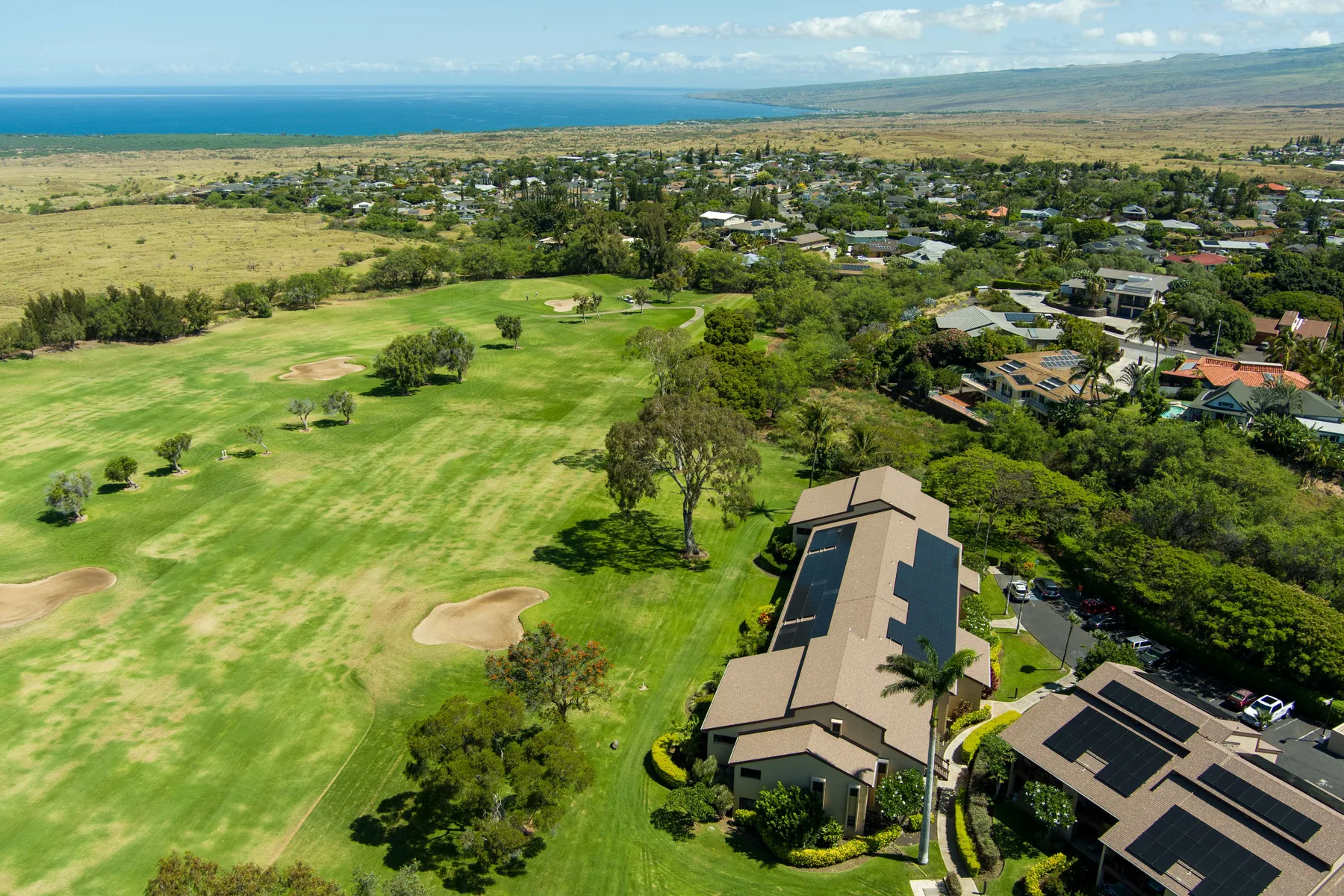 Aerial view of a golf course beside homes, with the ocean visible in the distance.