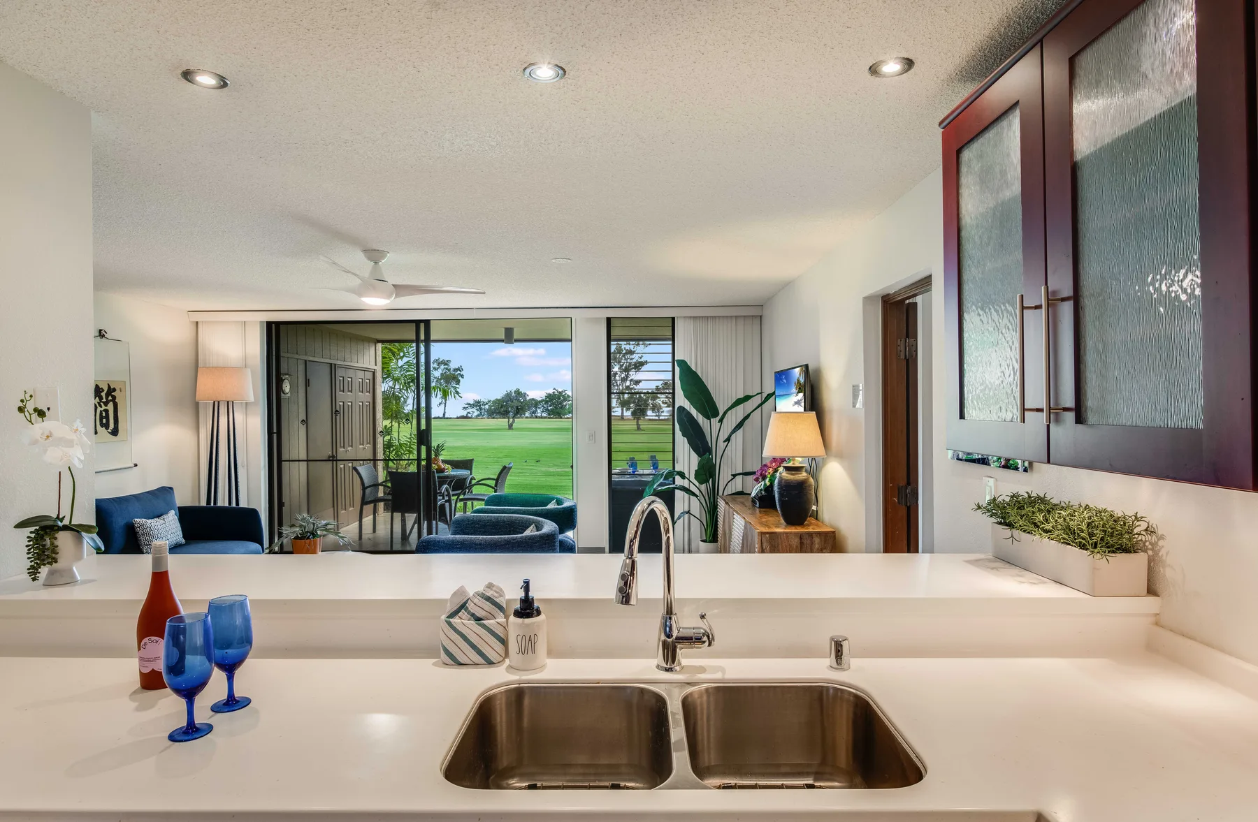 A bright kitchen counter with a double sink overlooks a living area and green landscape.
