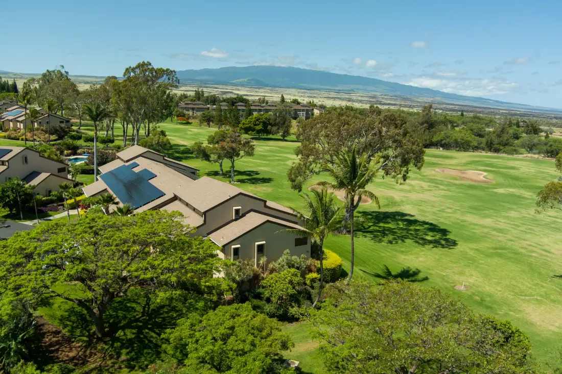 Aerial view of a landscaped golf course beside low buildings and distant mountains.