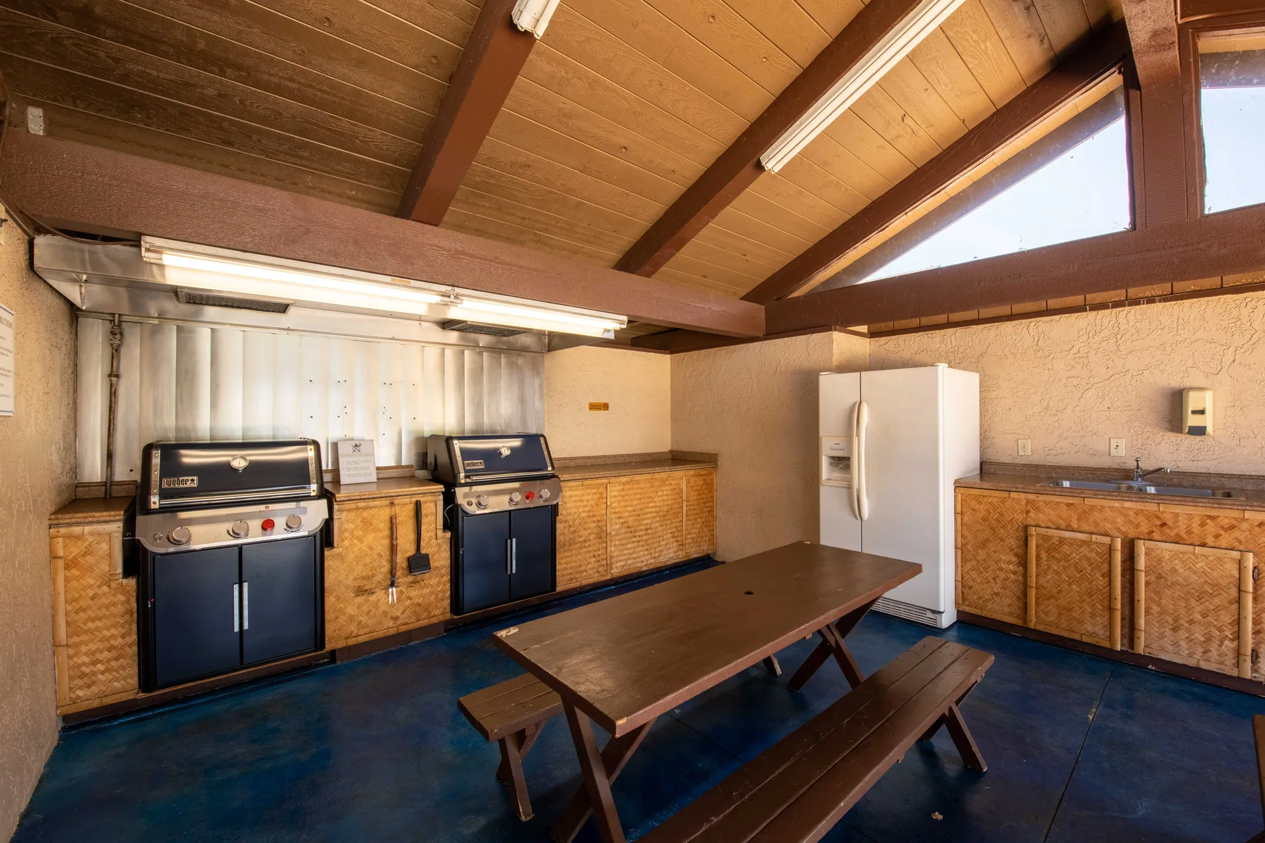 A covered barbecue area with grills, a refrigerator, and a picnic table.