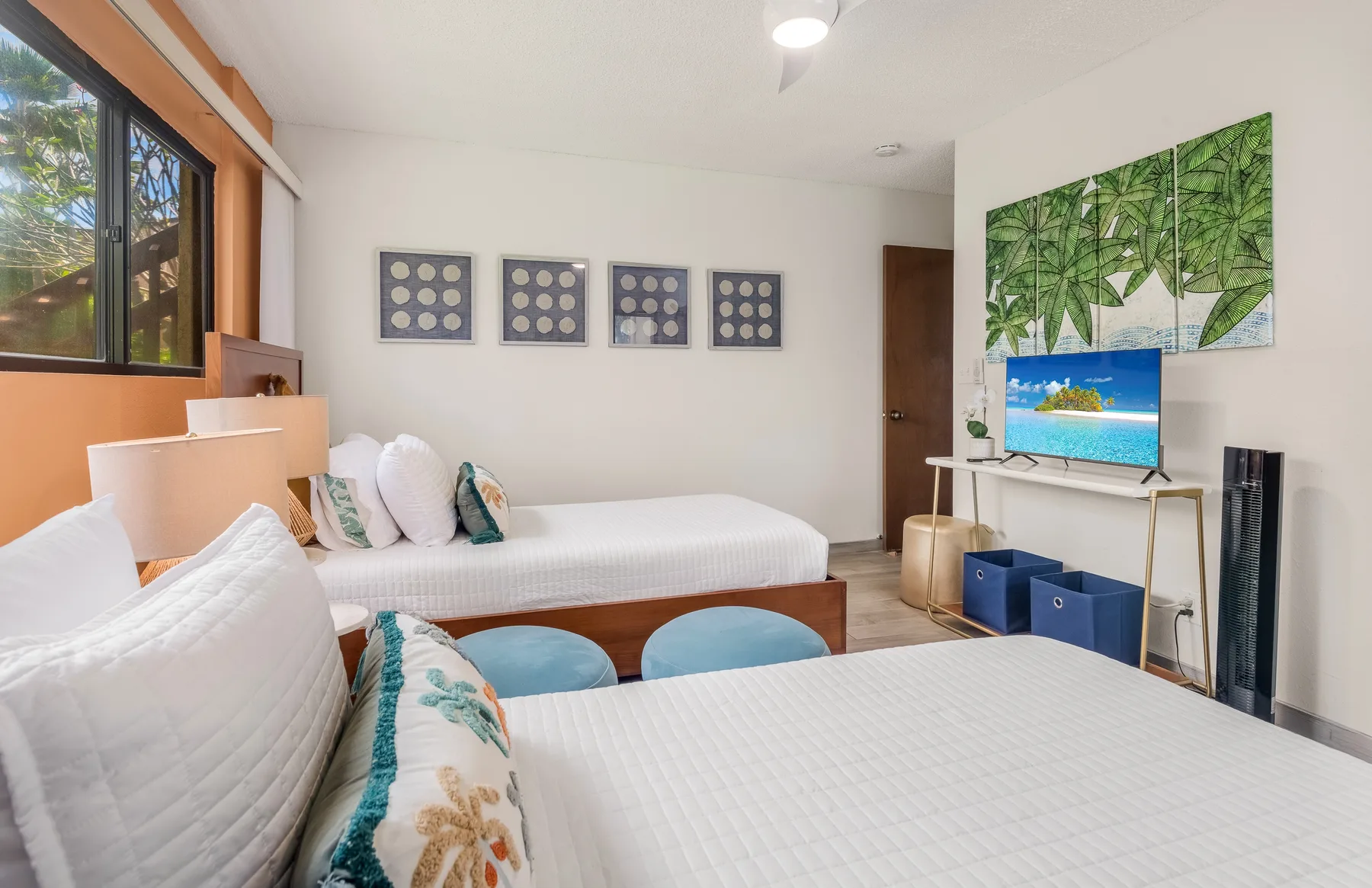 A bedroom with two beds, blue seating, and a television beneath leafy wall art.