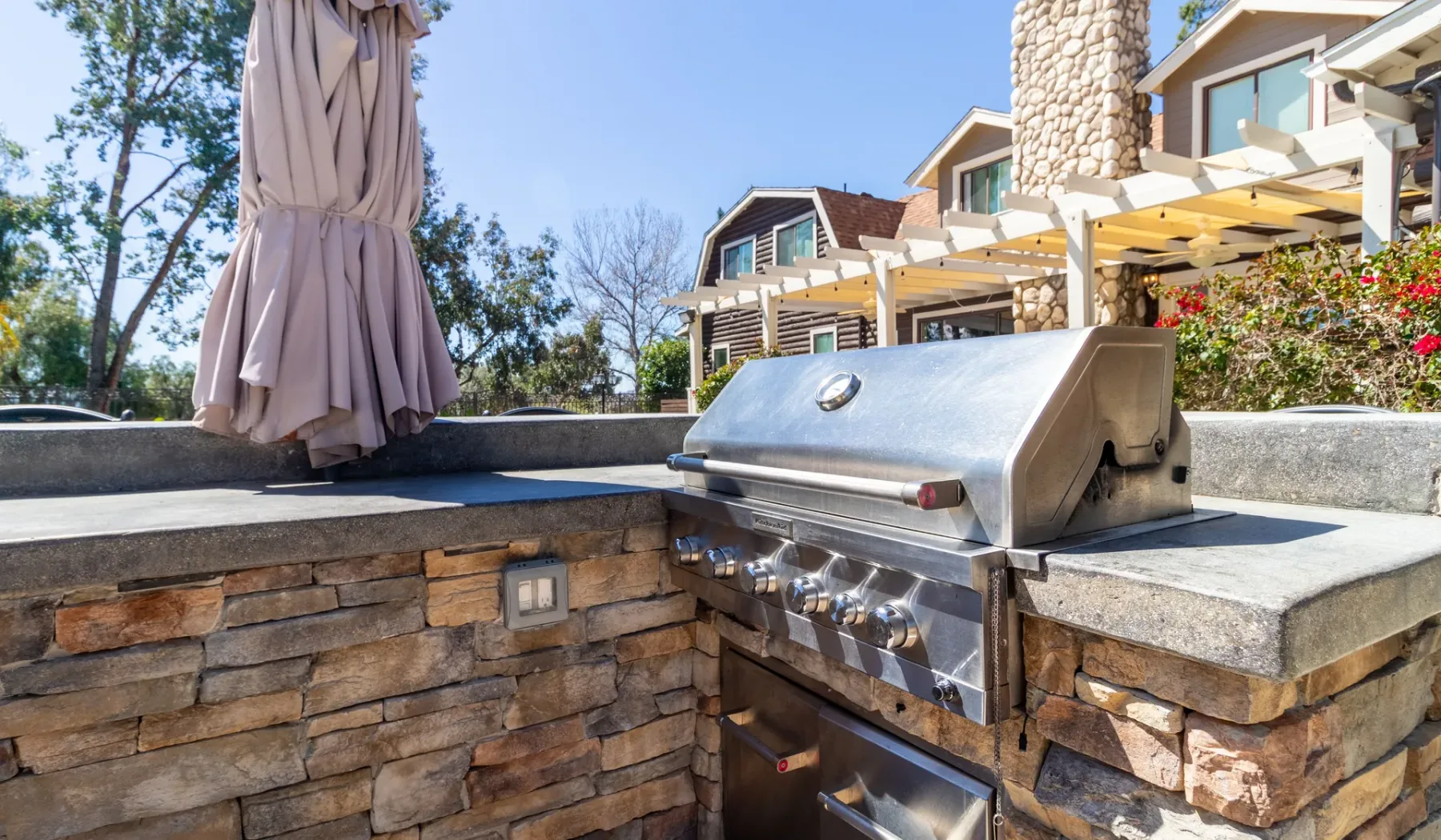 An outdoor stone counter features a stainless-steel grill and a closed patio umbrella.