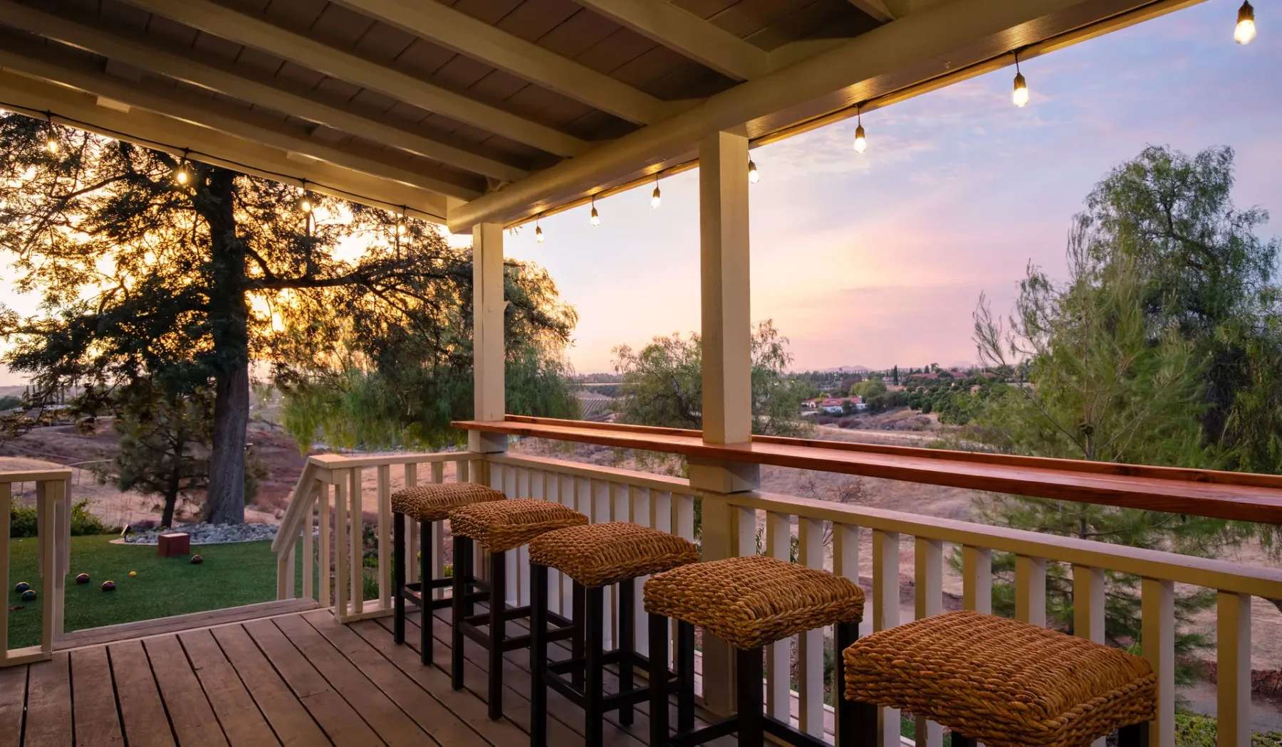 Una terraza de madera cubierta tiene asientos de barra y da a árboles al atardecer.