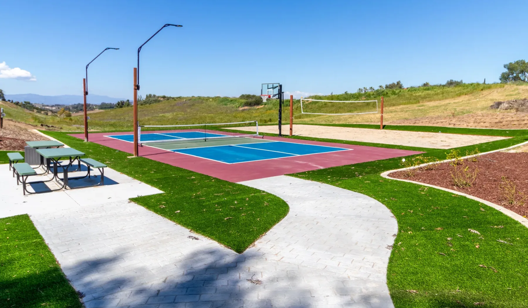An outdoor recreation court with basketball hoops, a volleyball net, and nearby picnic tables.