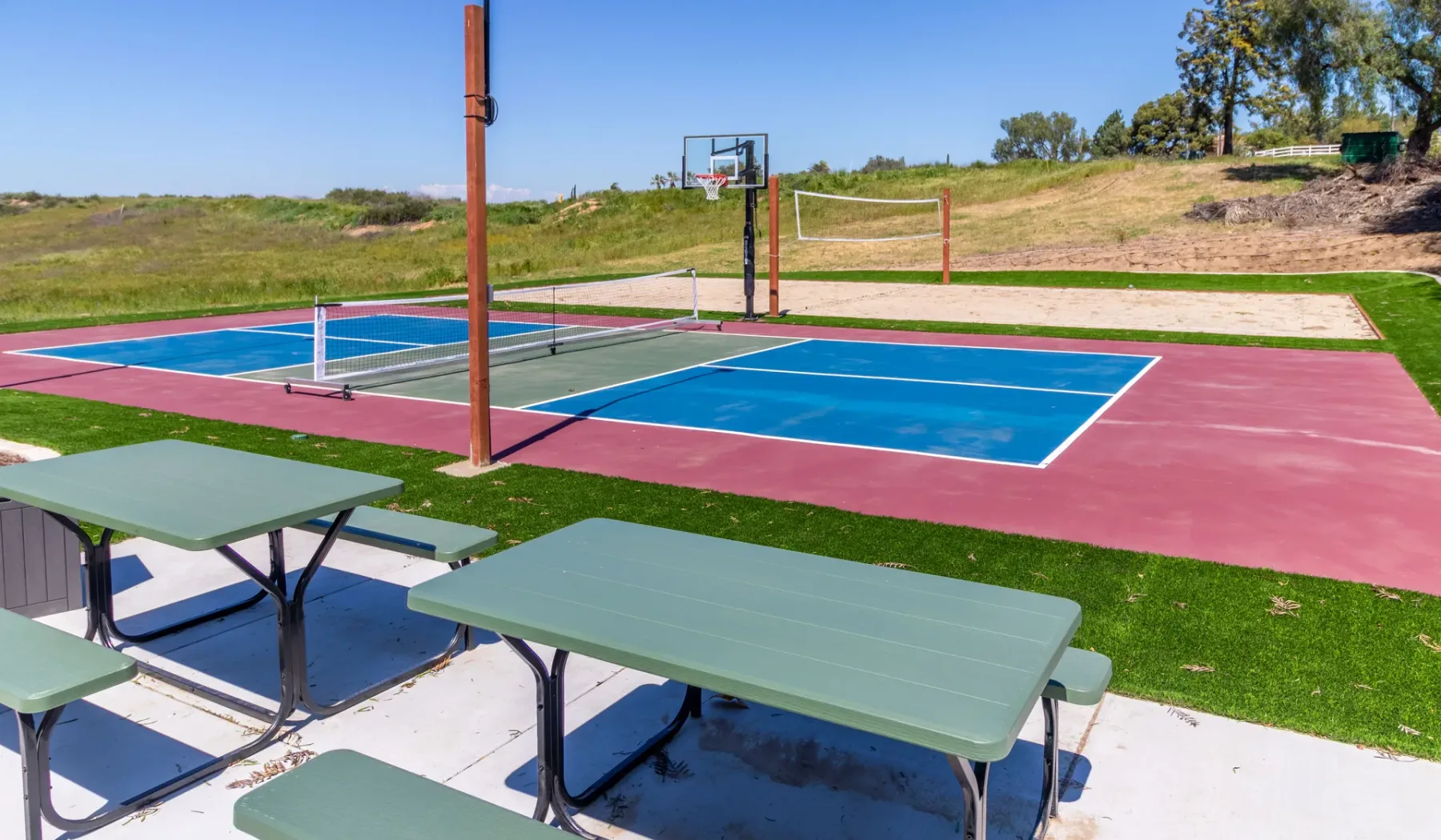 An outdoor sports court has basketball hoops, a net, and picnic tables nearby.