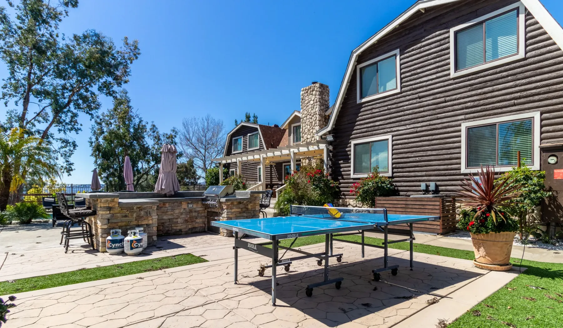 A paved outdoor patio beside a log-sided house features a table tennis table.