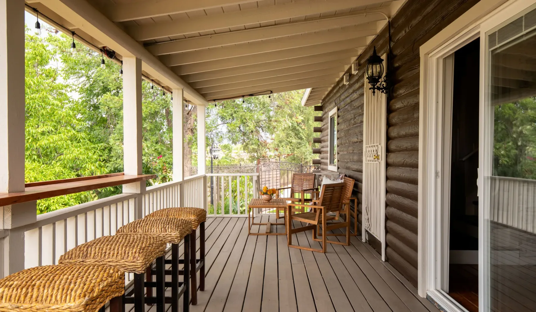 A covered wooden porch with seating and a railing overlooking green trees.