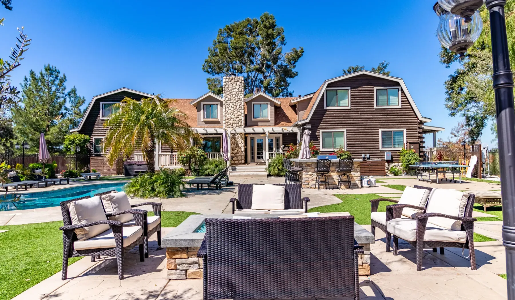 A landscaped outdoor patio with seating faces a large house and swimming pool.