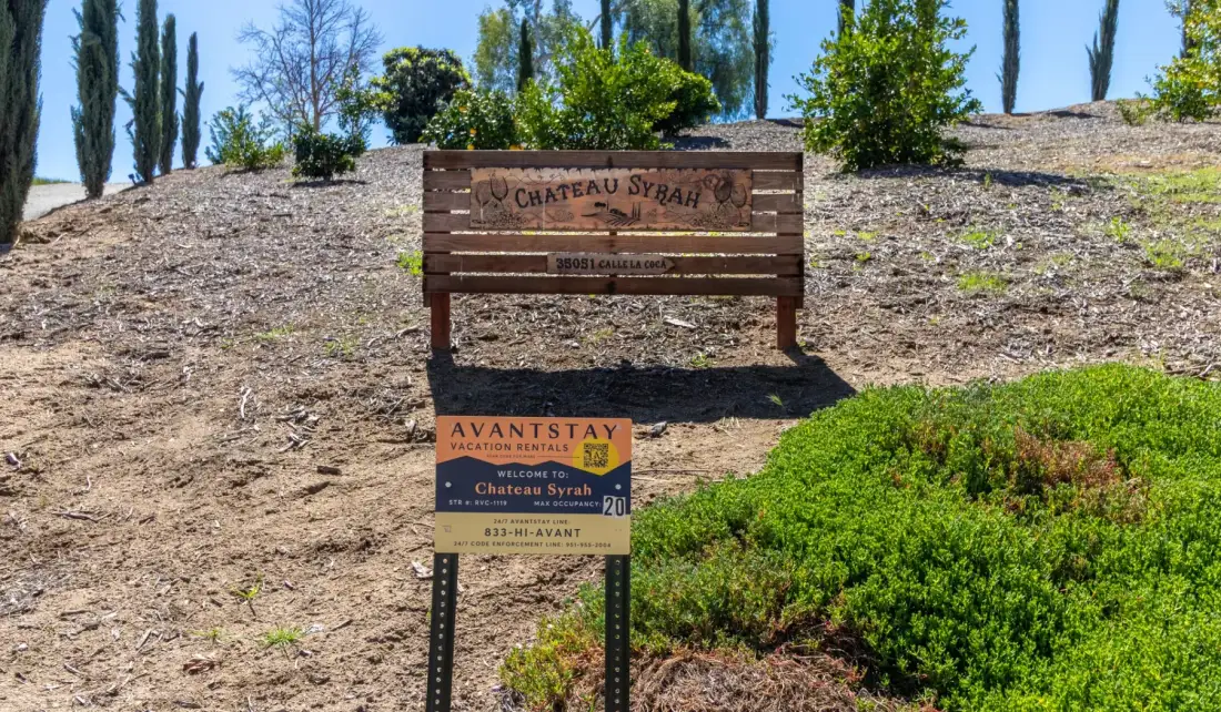 A wooden vineyard sign stands on a dry hillside among shrubs and tall trees.