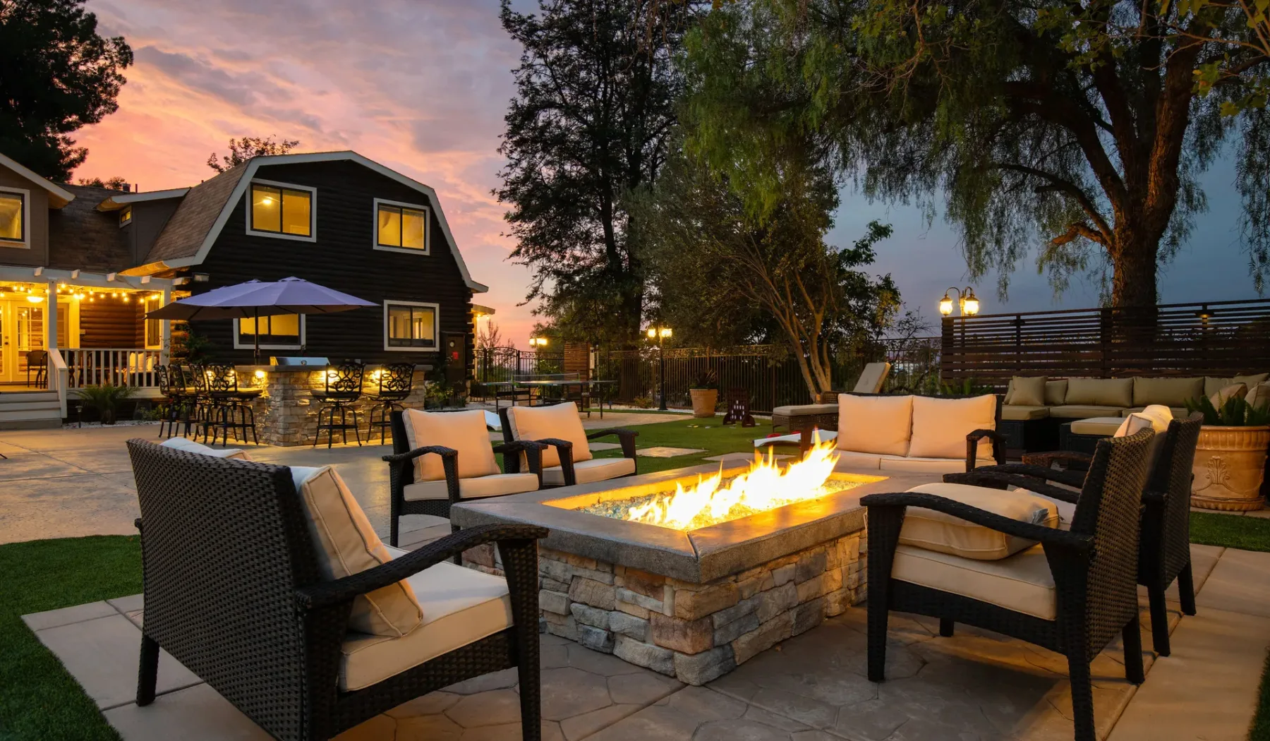 An outdoor patio seating area surrounds a lit stone fire pit at dusk.