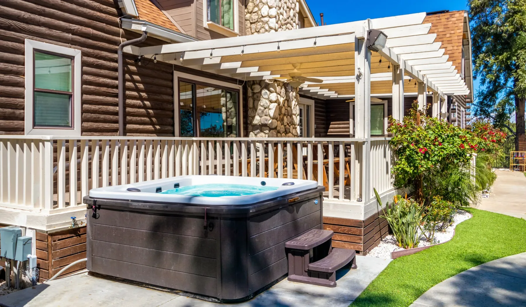 An outdoor hot tub sits beside a wooden house and covered patio.