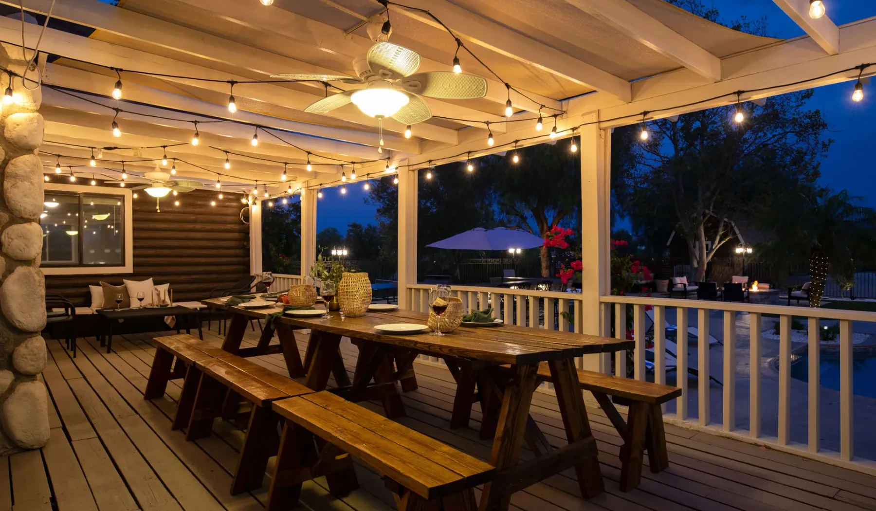 A covered outdoor deck with long wooden tables and warm string lights.