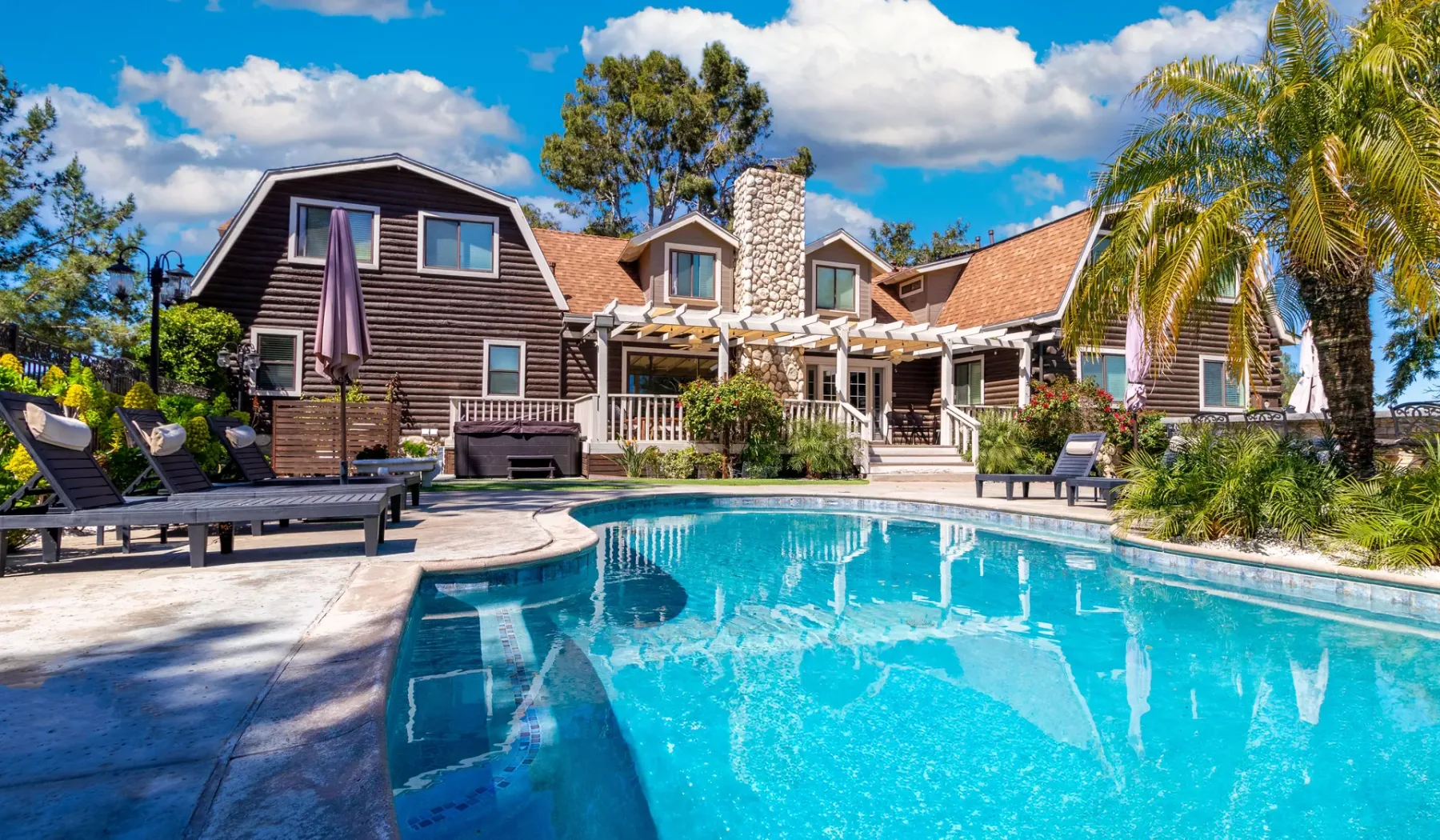 A swimming pool sits in front of a large house, surrounded by patio seating and palm trees.