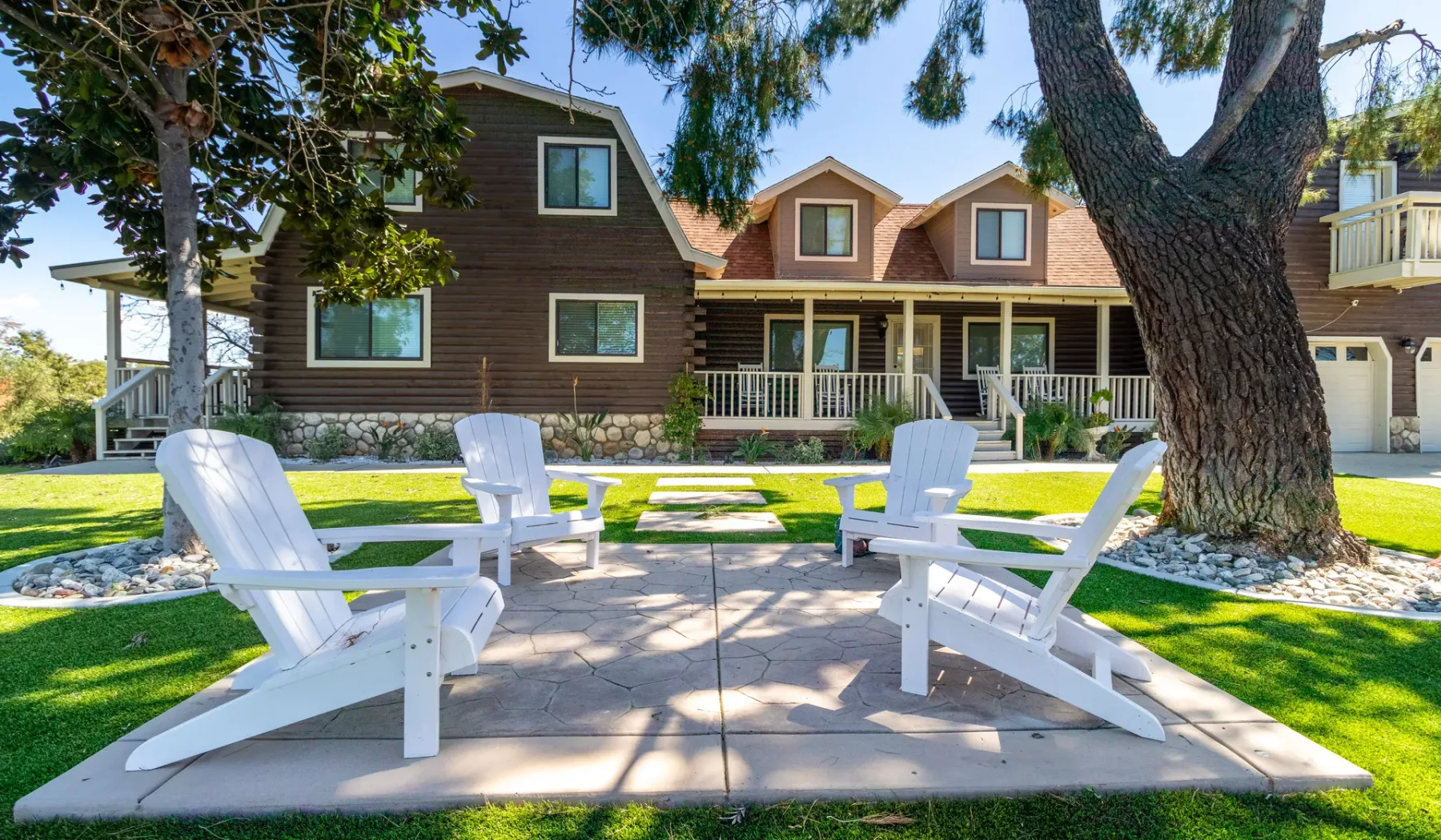 A landscaped outdoor patio has white seating beneath a large tree, with a lodge-style building behind it.
