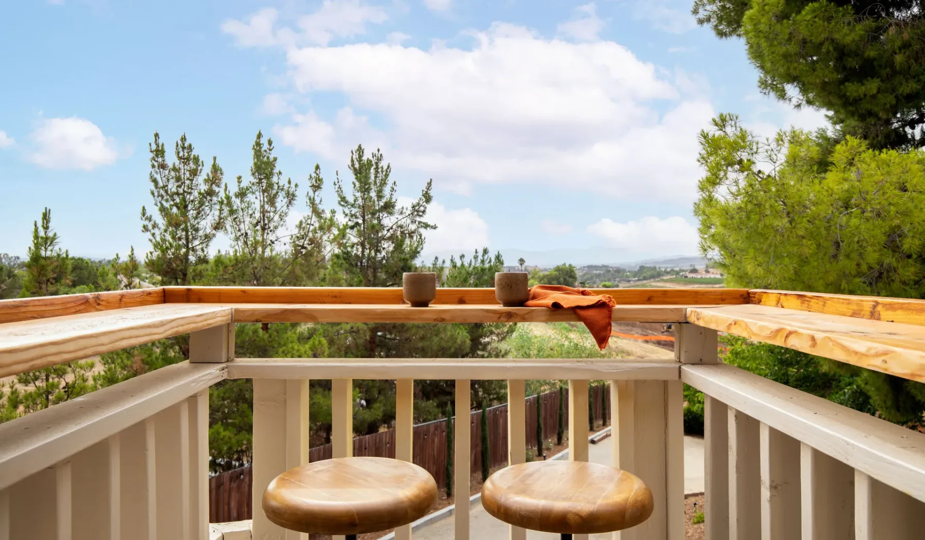 An outdoor wooden balcony with two round stools overlooking trees and open land.