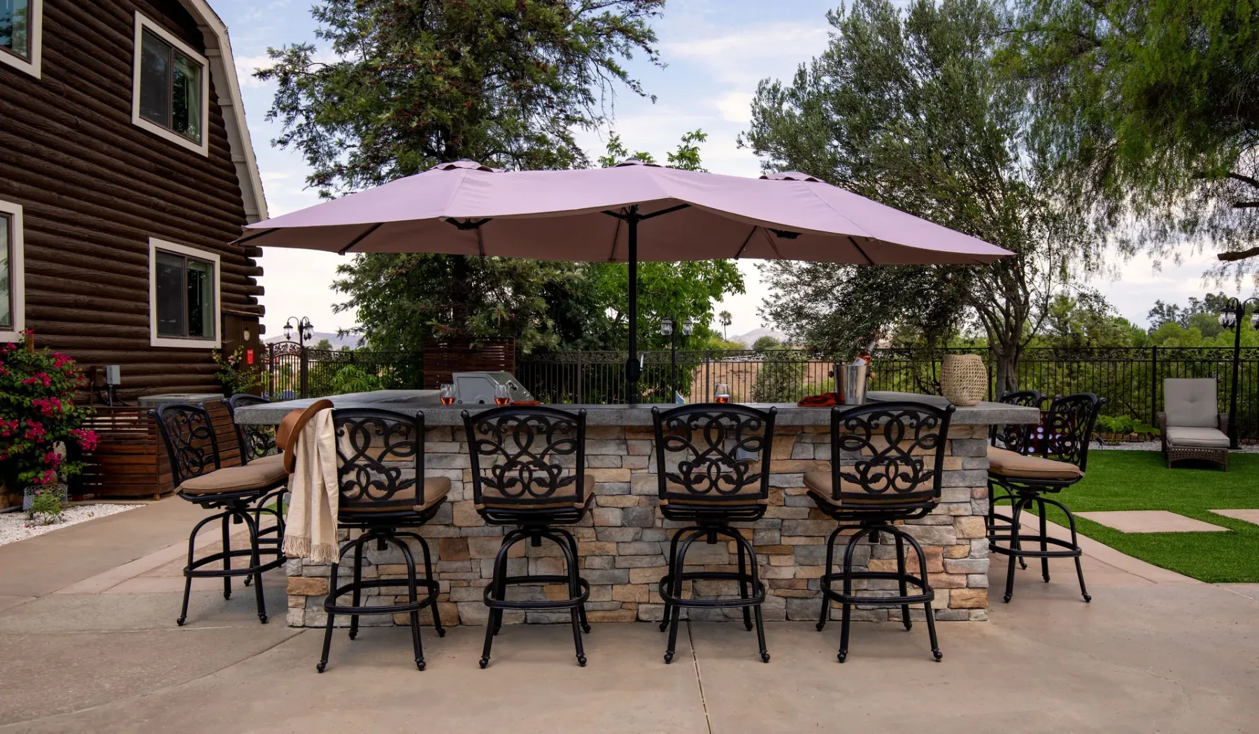 An outdoor bar counter with seating sits beneath a large umbrella on a patio.