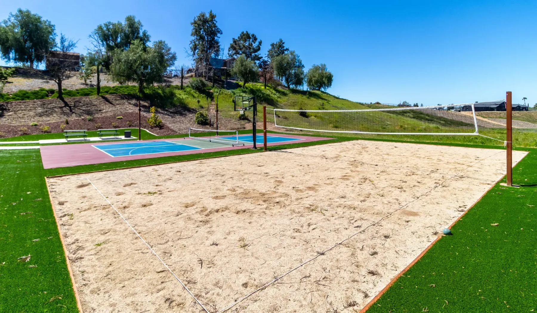 An outdoor sand volleyball court sits beside a colorful sports court on a sunny day.