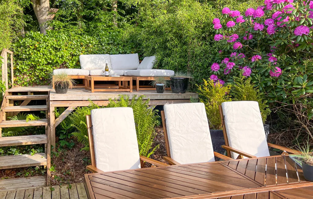 Un patio ajardinado con asientos de madera, sillones y arbustos en flor.