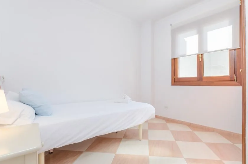 Un dormitorio luminoso con una cama blanca junto a una ventana con marco de madera.