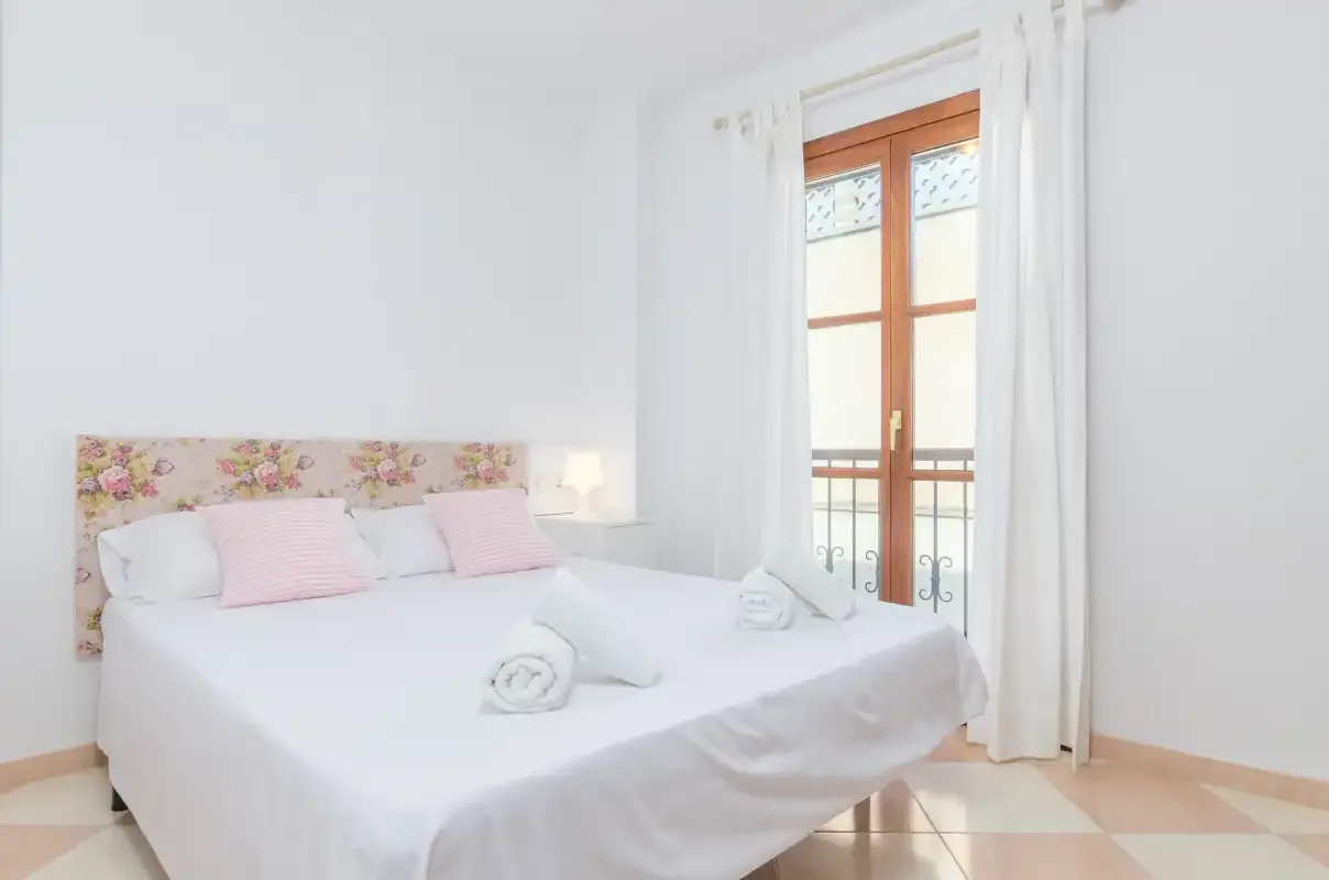 Un dormitorio luminoso con una cama blanca y una ventana con cortinas.