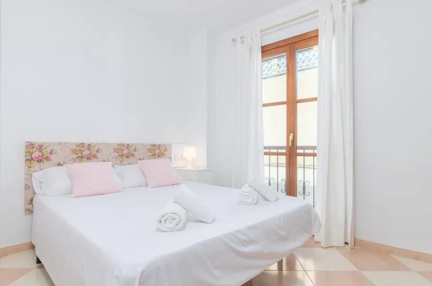 Un dormitorio luminoso con una cama blanca y una ventana alta enmarcada por cortinas.
