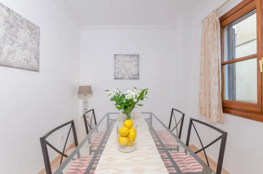 Un comedor con una mesa de vidrio, asientos y flores en un jarrón.