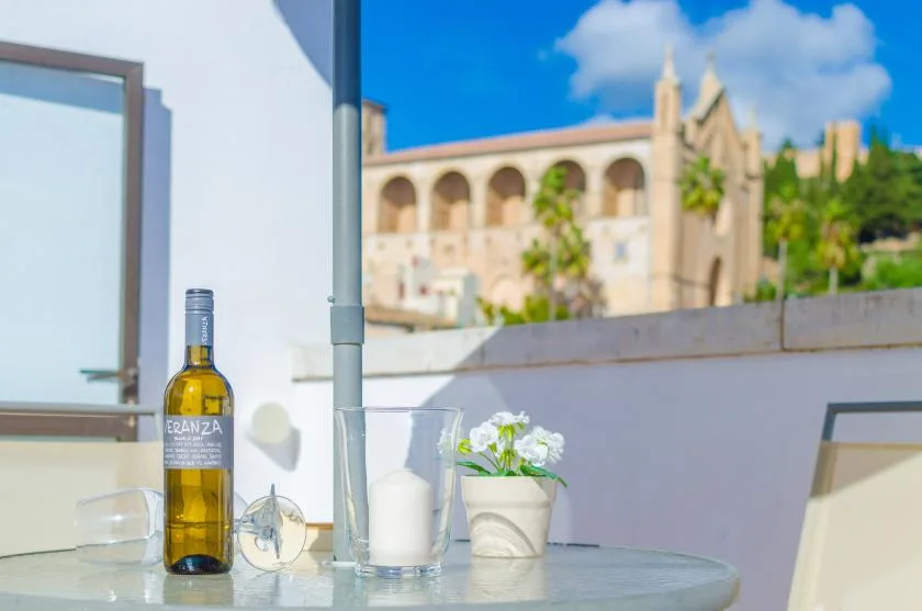 Una mesa en una terraza soleada tiene una botella, un vaso, una vela y flores en maceta.