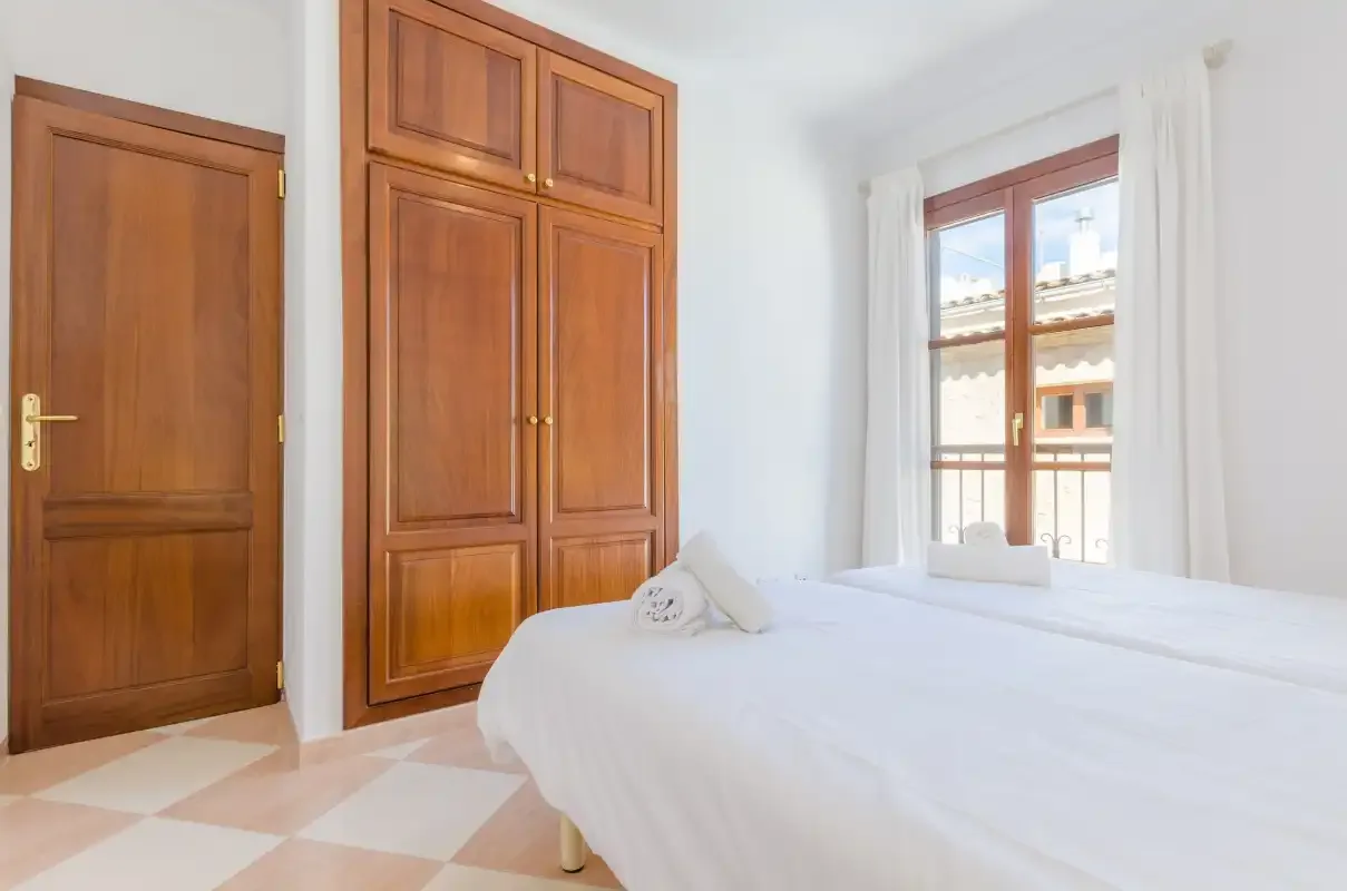 Un dormitorio luminoso con una cama blanca, un armario de madera y una ventana.