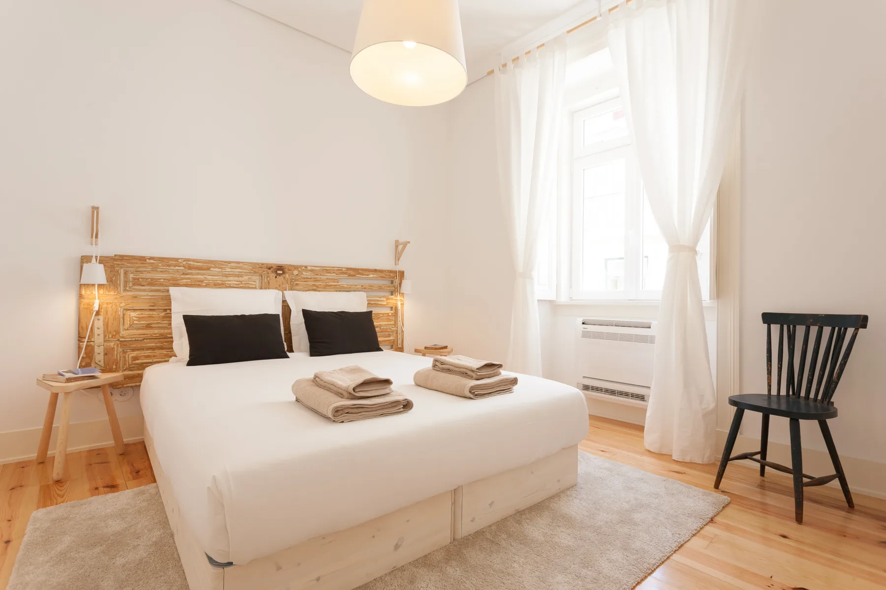 A bright bedroom with a white bed, wooden headboard, and a window with sheer curtains.