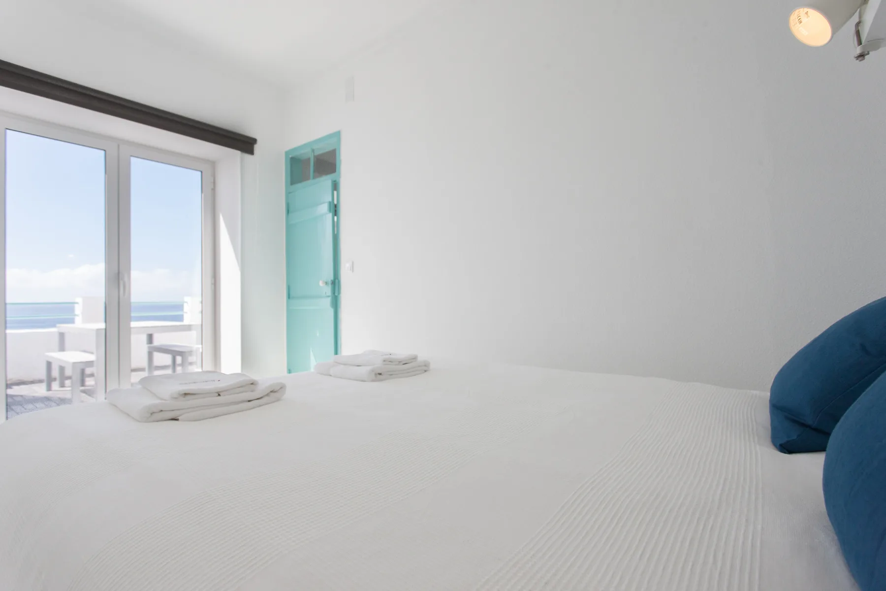 A bright bedroom with a white bed, turquoise door, and ocean view through glass doors.
