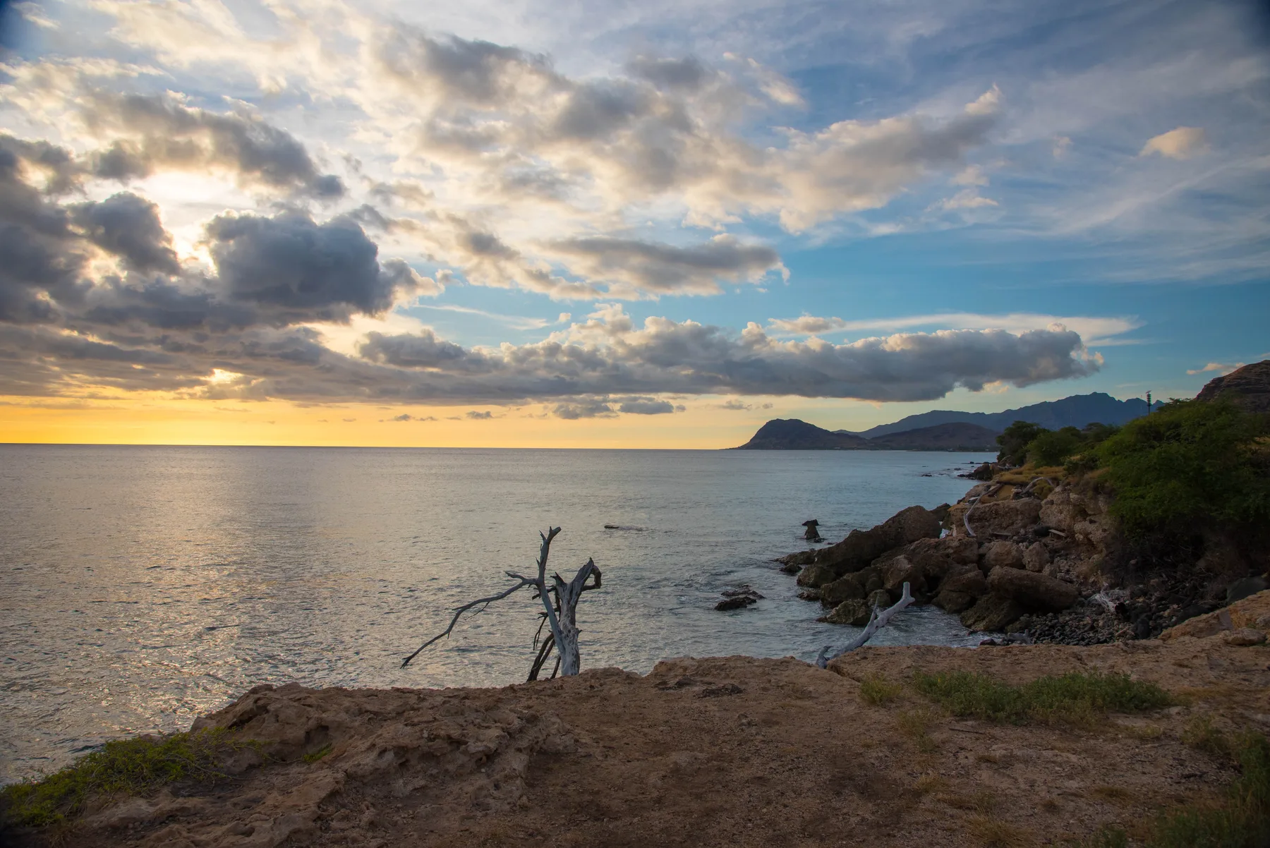 A rocky coastline and calm sea beneath a cloudy sunset sky.
