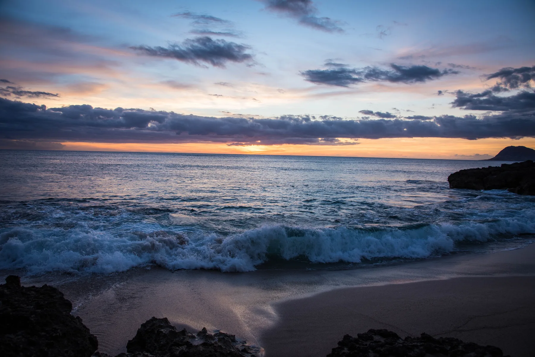 Ocean waves reach a sandy beach beneath a colorful sunset sky.