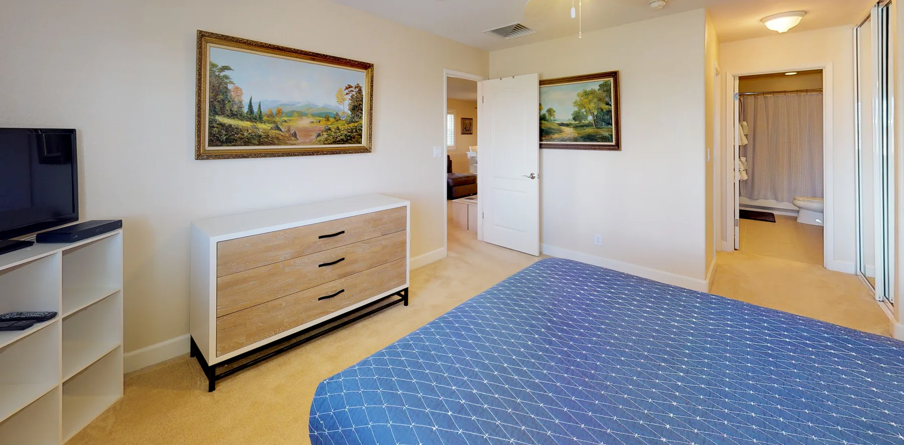 A bedroom with a blue patterned bedspread, dresser, and framed landscape paintings.