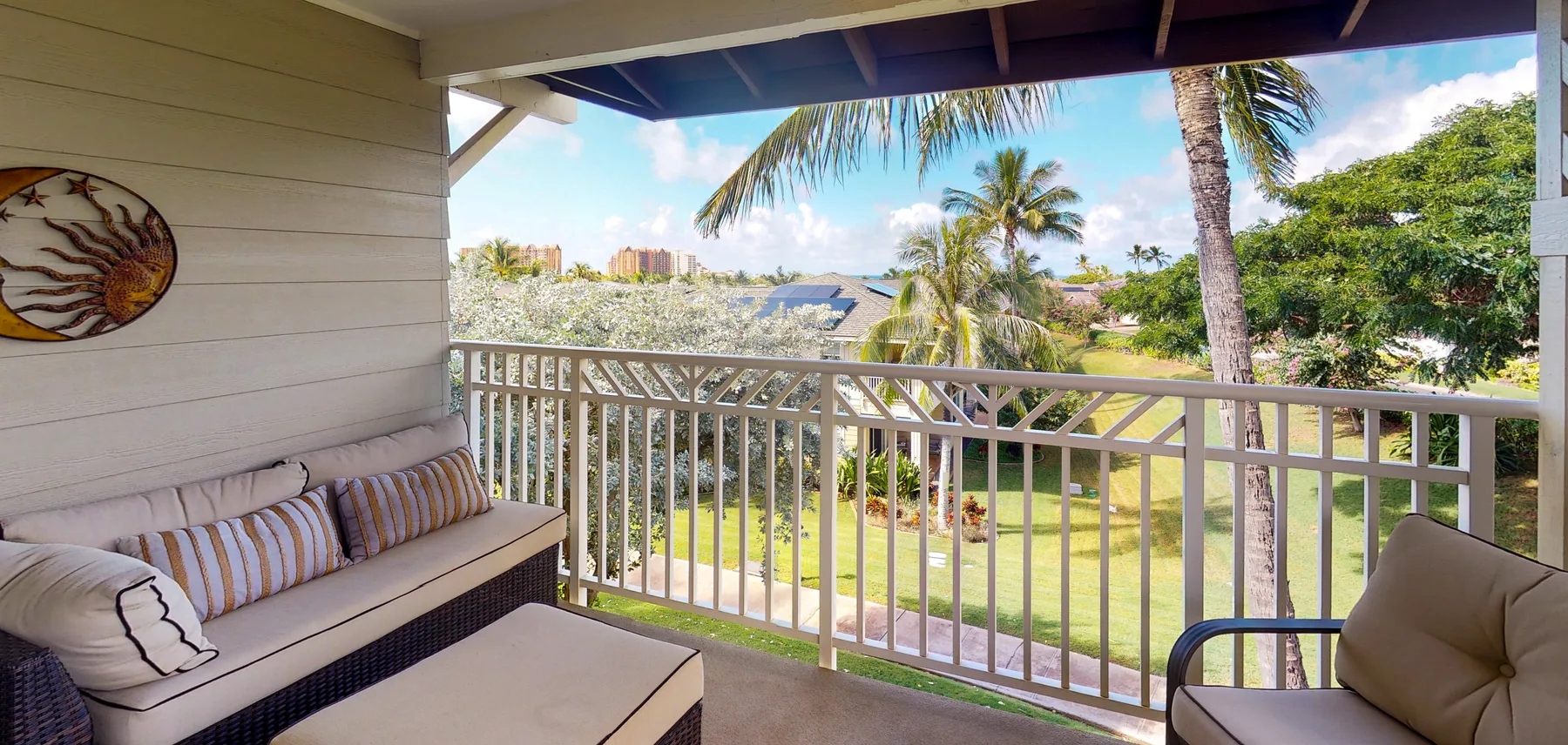 A covered balcony with cushioned seating overlooks a green garden and palm trees.