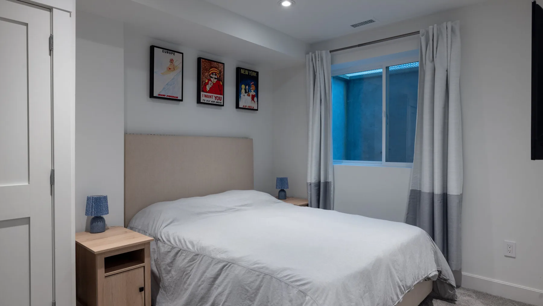 Un dormitorio con una cama, mesas de noche, cuadros enmarcados y una ventana con cortinas.