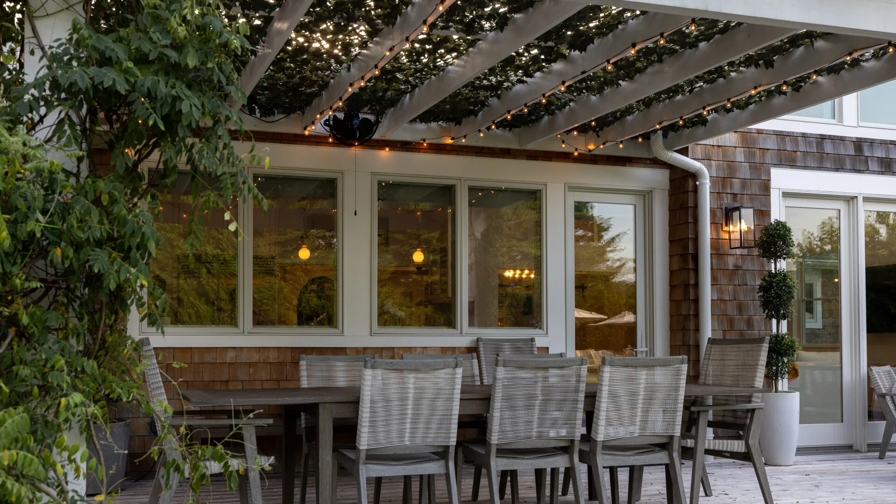 Una zona de comedor al aire libre está bajo una pérgola con plantas, junto a grandes ventanas.