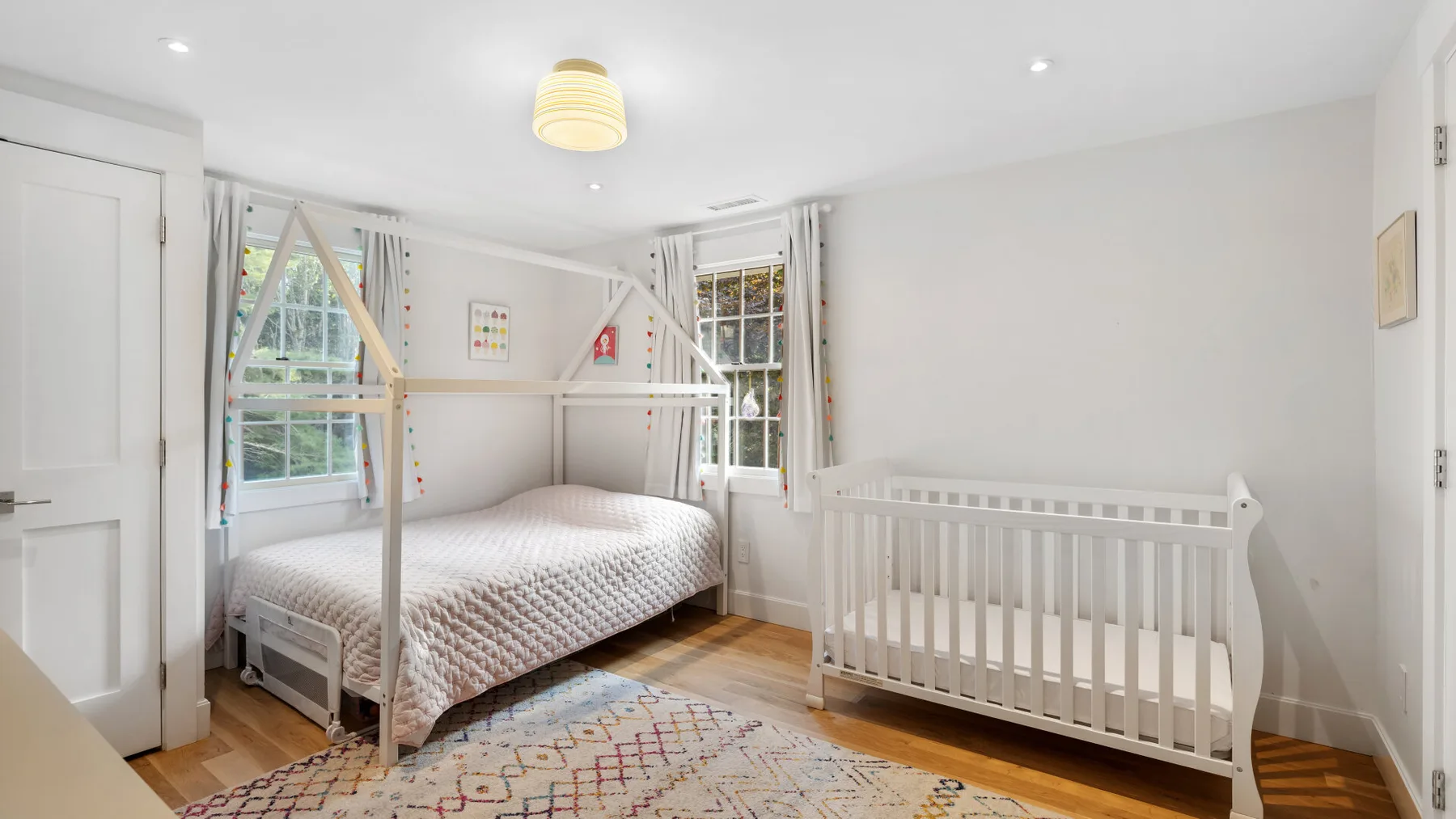 Un dormitorio infantil luminoso con una cama y una cuna blanca junto a las ventanas.