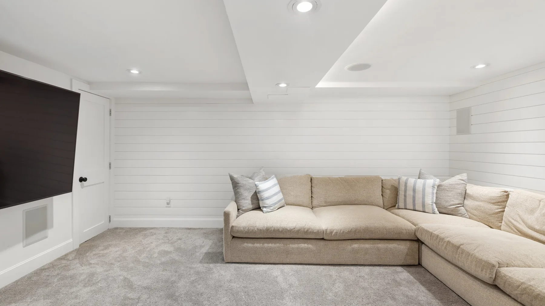 Una sala luminosa con un sofá seccional beige y un televisor en la pared.